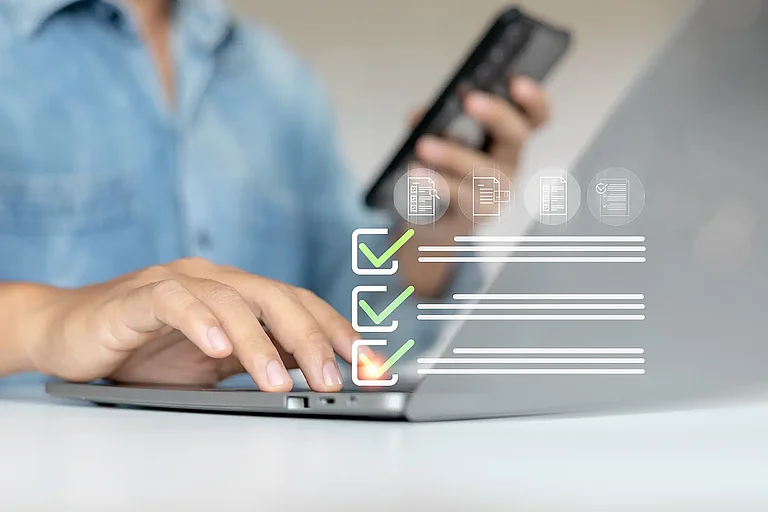 Eine Person verwendet einen Laptop und hält ein Smartphone, während sie mit einer virtuellen Checkliste interagiert. Konzept der digitalen Aufgabenverwaltung, Formularübergabe und Produktivitätstools.