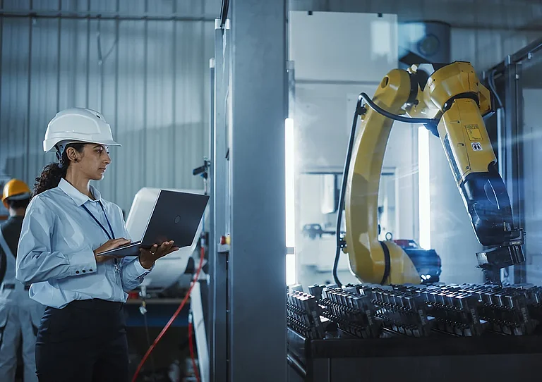 Eine Wirtschaftsingenieurin bei der Arbeit. Eine hispanische Frau überwacht mithilfe eines Laptops einen Roboterarm in einer Hightech-Metallfabrik, um eine präzise Automatisierung und Effizienz im Produktionsprozess sicherzustellen.