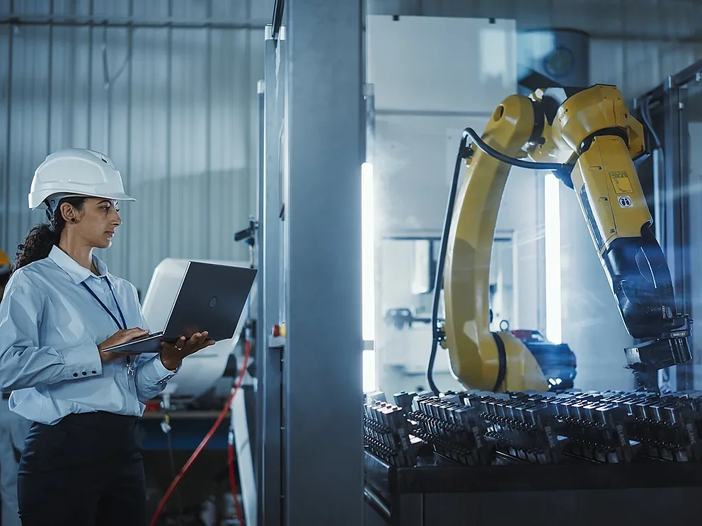 Eine Wirtschaftsingenieurin bei der Arbeit. Eine hispanische Frau überwacht mithilfe eines Laptops einen Roboterarm in einer Hightech-Metallfabrik, um eine präzise Automatisierung und Effizienz im Produktionsprozess sicherzustellen.