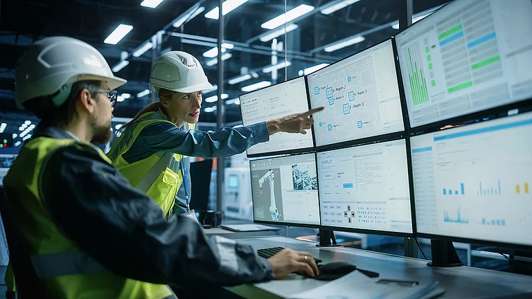 Ein weißer Prozessingenieur arbeitet an einem Arbeitsplatz mit mehreren Monitoren und unterhält sich mit einer Qualitätskontrolleurin in der Elektronikfabrik. Kollegen überwachen eine automatisierte Montagelinie, die mit Roboterarmen ausgestattet ist.