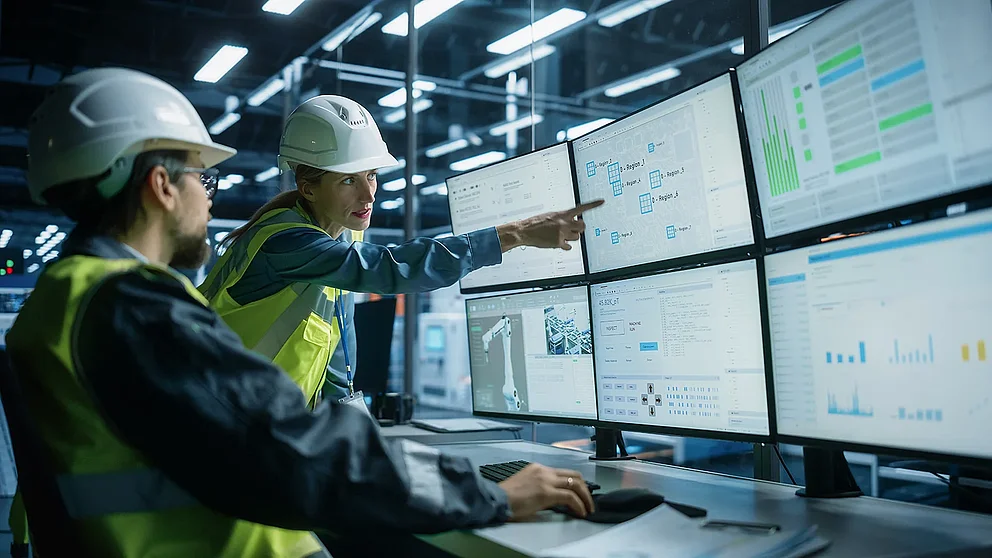 Ein weißer Prozessingenieur arbeitet an einem Arbeitsplatz mit mehreren Monitoren und unterhält sich mit einer Qualitätskontrolleurin in der Elektronikfabrik. Kollegen überwachen eine automatisierte Montagelinie, die mit Roboterarmen ausgestattet ist.